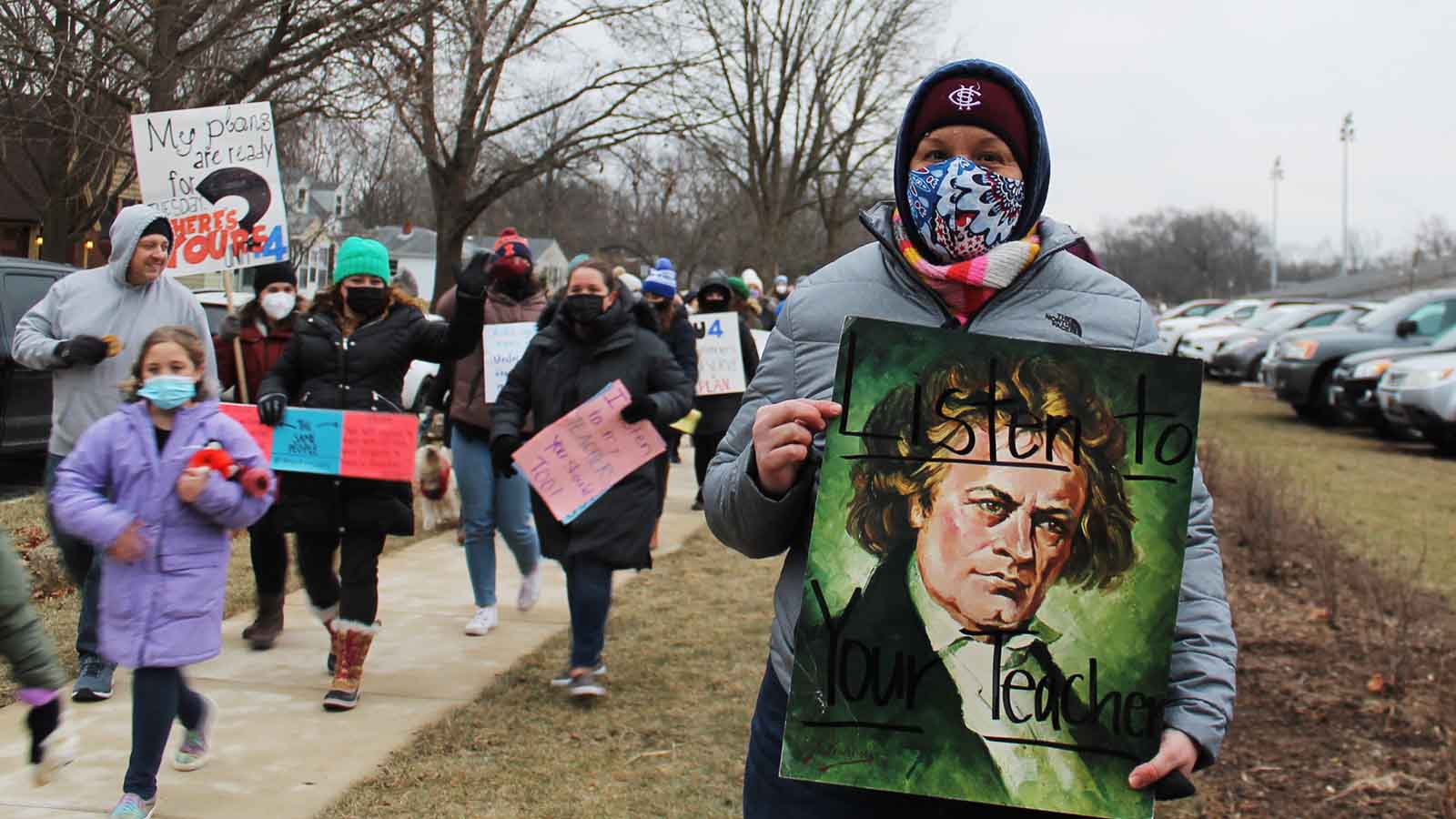 A woman with a face mask holds a sign that says, "Listen to your teacher."