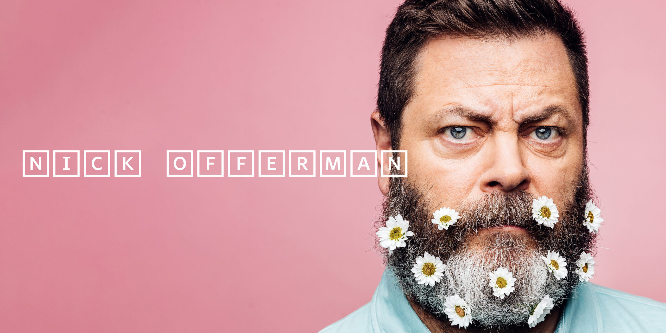 A headshot of Nick Offerman, a dark-haired man, with small, white flowers stuck into his graying beard sits on a pink backdrop.
