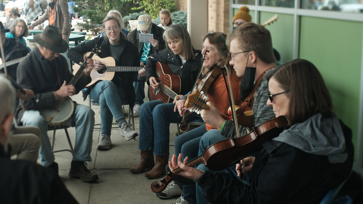 A large group of people sit in folding chairs on a concrete patio, playing a mixture of banjos, fiddles and guitars.