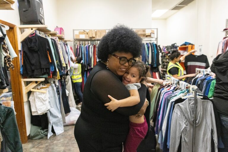 A woman is wearing a black shirt and glasses. With a smile on her face, the woman is hugging a little girl in a clothing store.