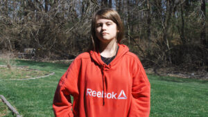 A young person with long, light brown hair stands outside. They are wearing an oversized, red, Reebok hoodie.