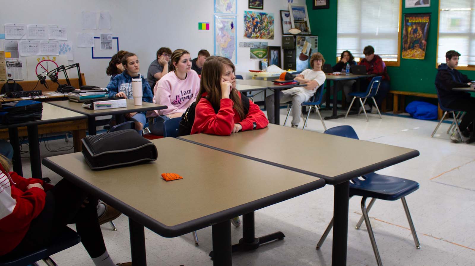 Eleven students sit around tables in a classroom with one green wall and one white wall. There are posters, maps and printed sheets on the walls, and there is podcasting equipment in one back corner. The student in the foreground has long, light brown hair and a big red hoodie.