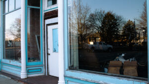 The storefront is painted blue and white. Large windows reflect trees, American flags and a truck. Inside, a pile of banners and other apparent junk is visible.