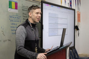 A white teacher with a short beard and glasses on top of his head speaks at a podium. There is a white board and a Smartboard behind him.