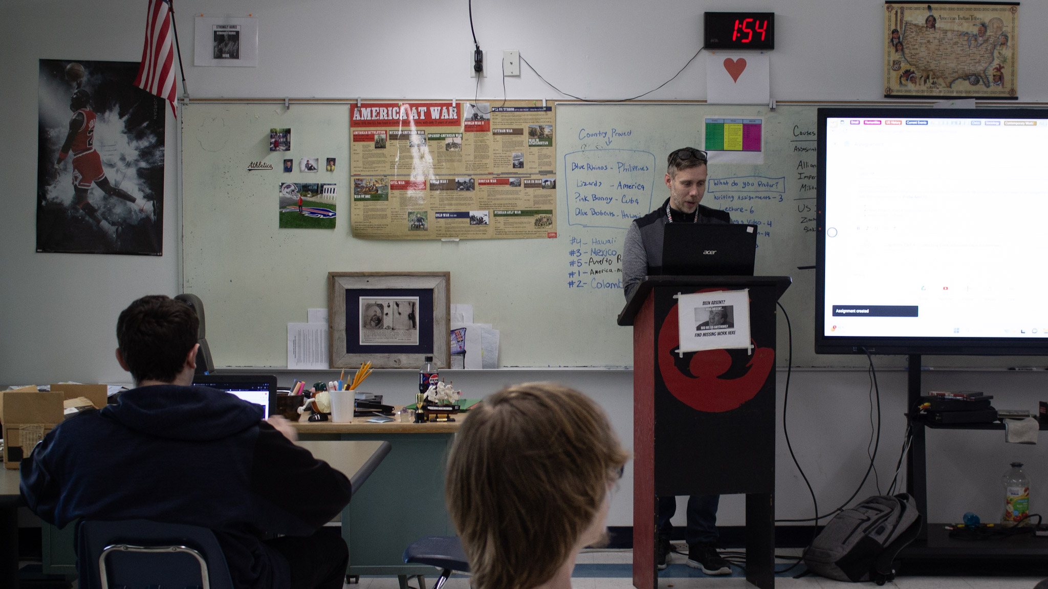 A white man stands behind a podium in a classroom. There is a SmartBoard and a white board behind him. The backs of two students' heads are in the foreground. A clock over the whiteboard says 1:54.