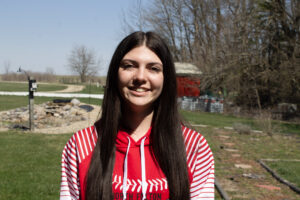 A white girl in a white and red sweatshirt and straight brown hair stands outside. The grass behind her is mowed, and there is a shed and some stone landscaping.