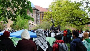 Six protesters are in the foreground with their backs to the camera. One protester wears a black and white, checkered scarf for Palestine. Blue and green tents, green leaves and campus buildings are in the background.