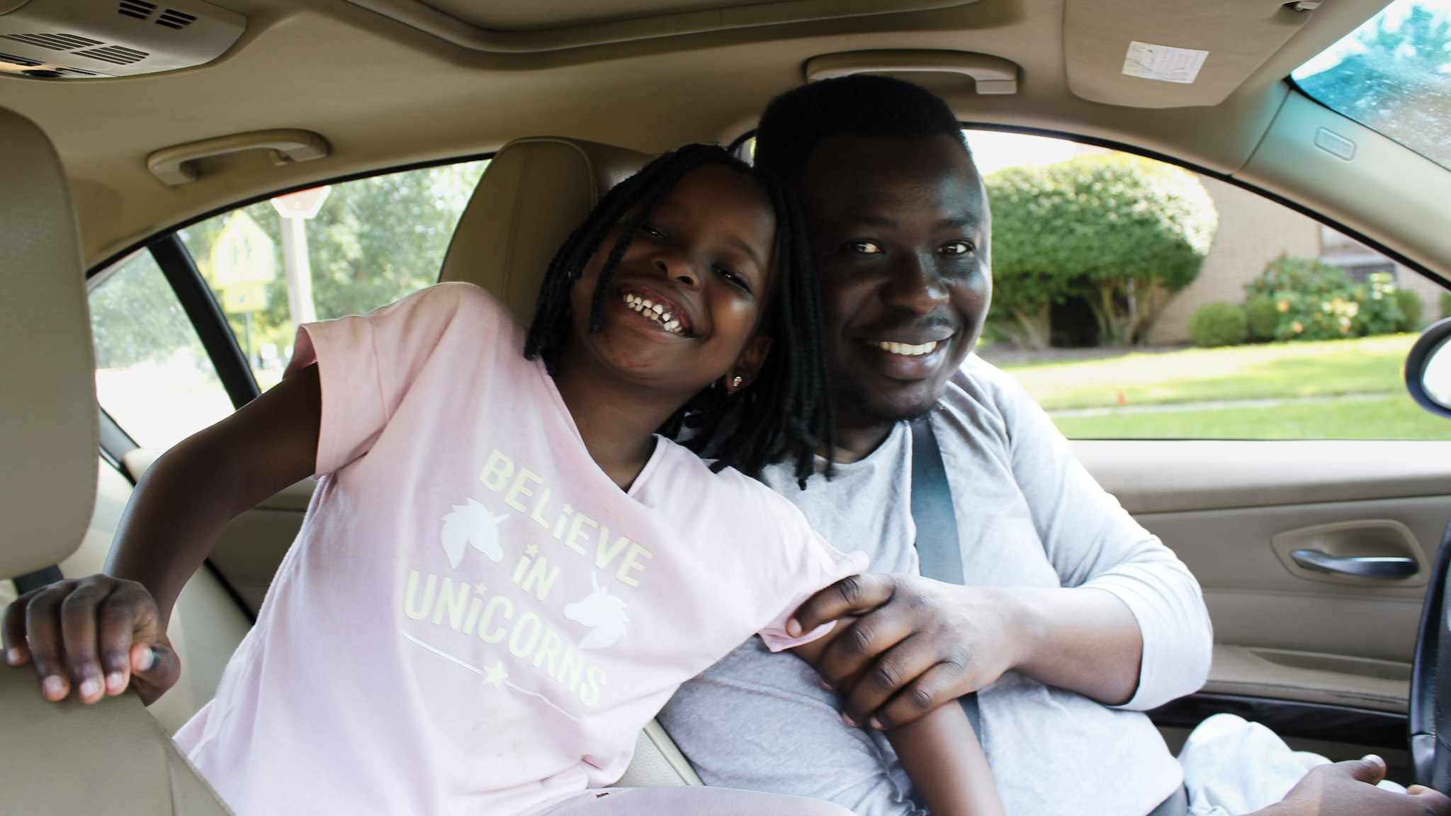 A 6-year-old grins at the camera in a pink t-shirt while her father holds her arm. Both are in a car.