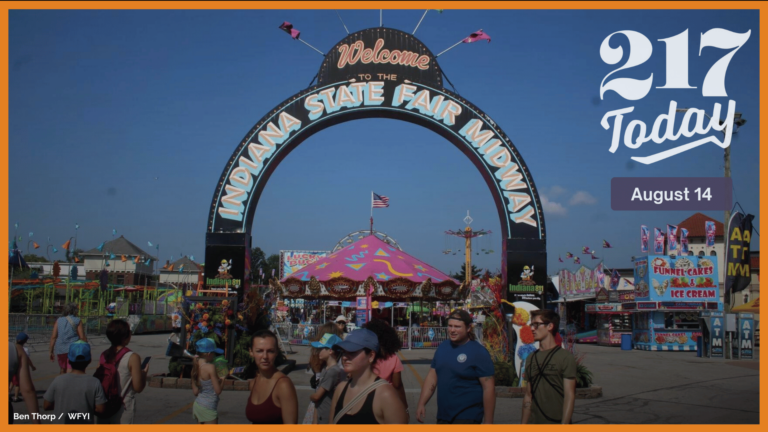 People are walking in front of a sign that says "Welcome to the Indiana State Fair Midway." Carnival rides and games can be seen in the background of the photo.