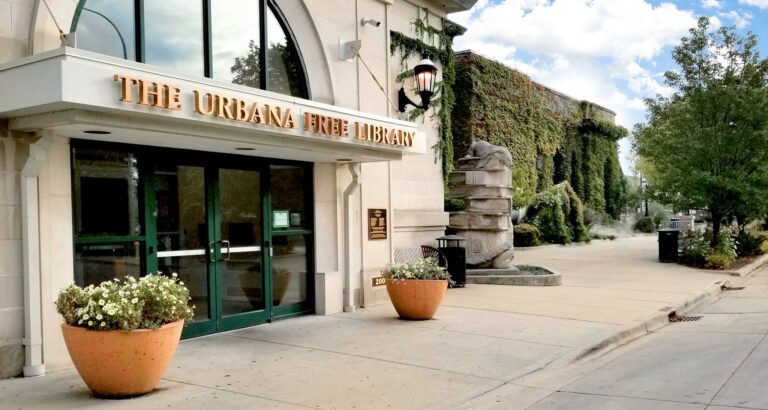 A courtyard in the background of a building with a sign reading "The Urbana Free Library"