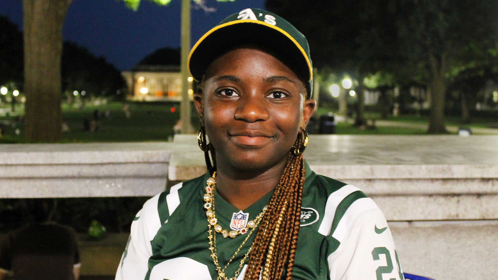 A Black student with braids and a green jersey smiles at the camera. It's night and the University of Illinois Quad is behind her.