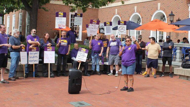 A woman holds a microphone. People with SEIU shirts and signs stand in the background.