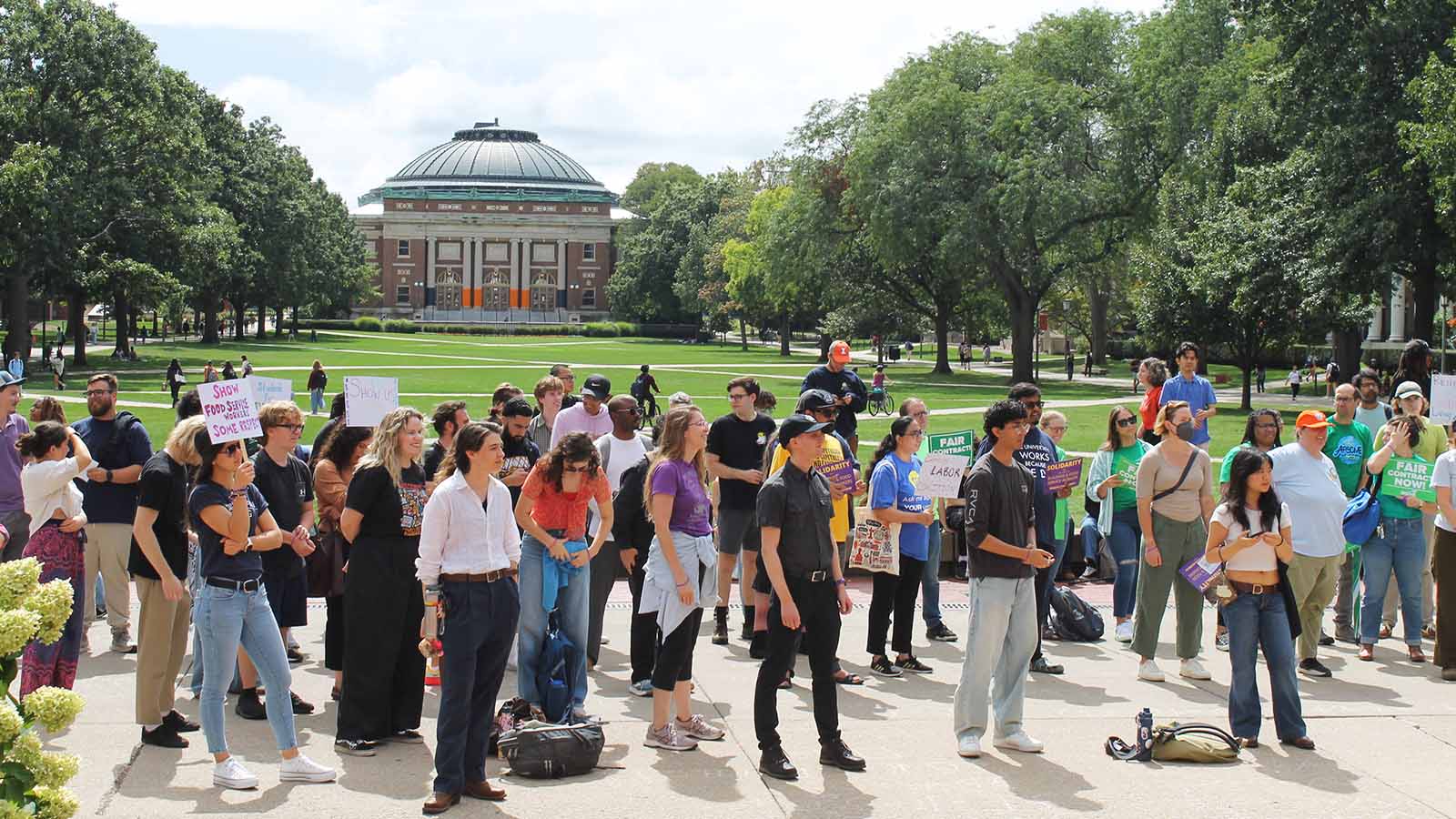 Some in the crowd have signs. Behind them is green lawn, trees and the iconic dome of Follinger Hall.