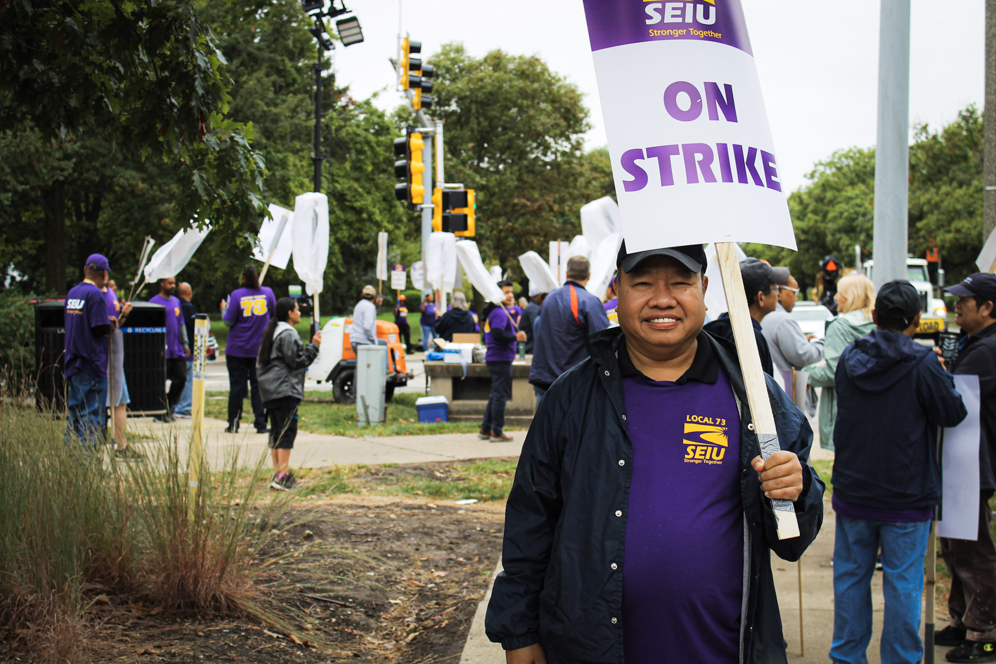 A Vietnamese dining hall worker holds a "On Strike" sign. Other workers are behind him on the sidewalk at an intersection.