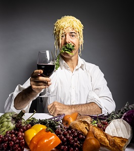 A man in a white shirt with noodles on his head and greens in his mouth holds up a glass of wine with a spread of food in front of him.