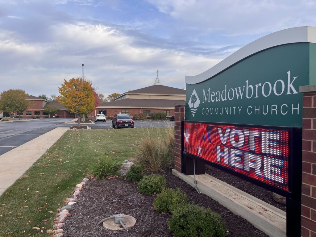 A sign for the Meadowbrook Community Church that says "vote here" below next to a parking lot.