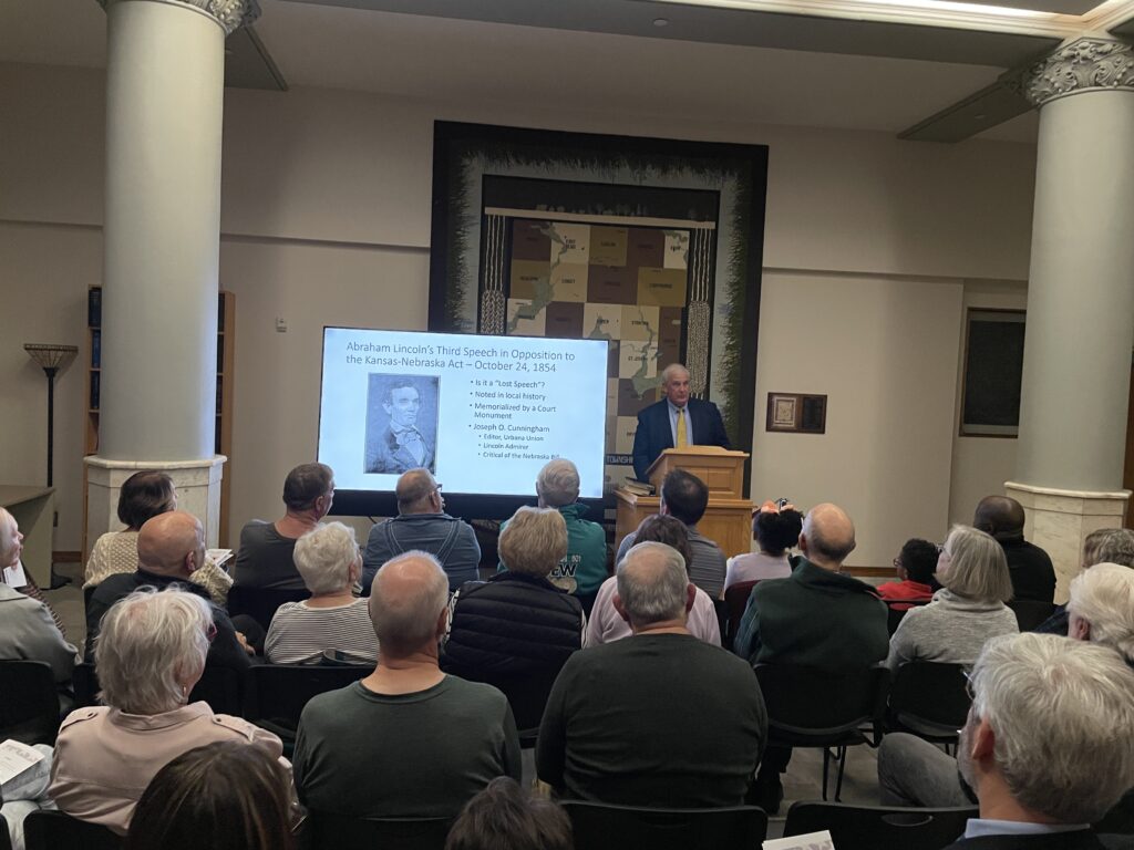 A man stands before a crowd next to a slideshow displaying information about Abraham Lincoln's visit to Urbana.