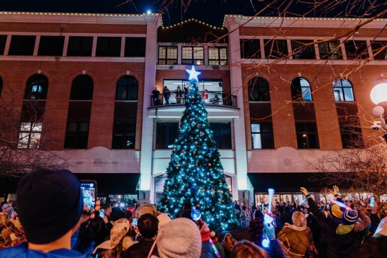 People surround a lit up tree in front of a building.