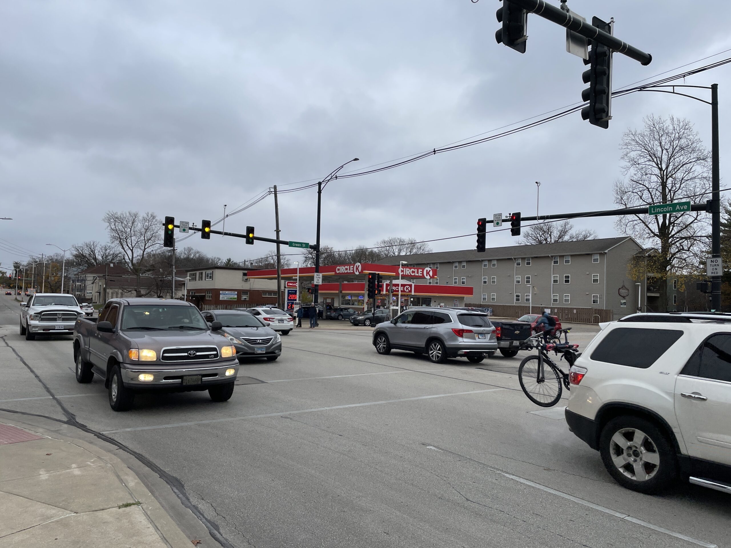 Several car and truck drivers head south on South Lincoln Avenue adjacent to a gas station. A cyclist is also visible behind the cars.