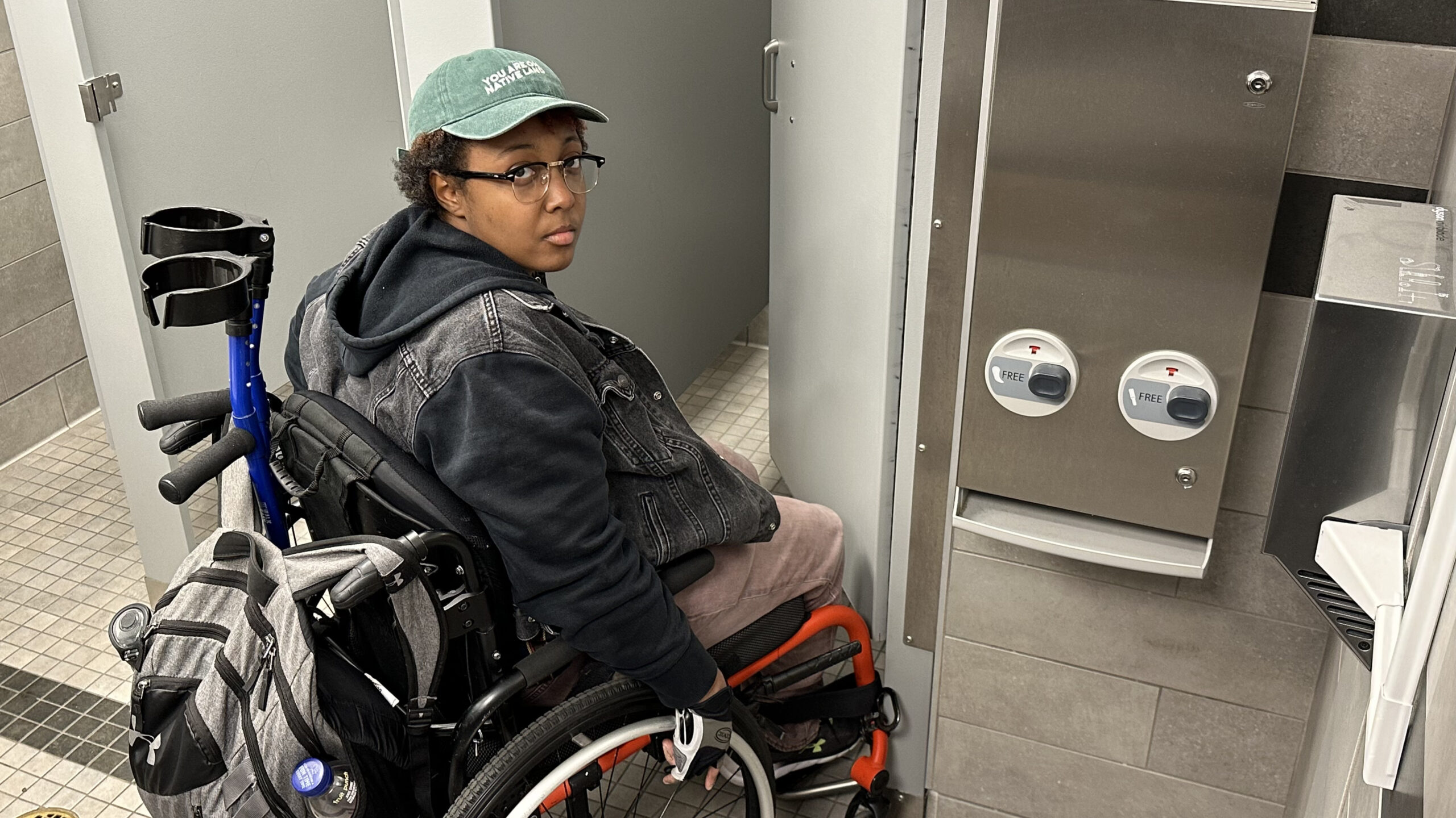 a person in a wheelchair in a bathroom. the person is wearing a green hat, a jacket and has a backpack attached to the back of the wheelchair.