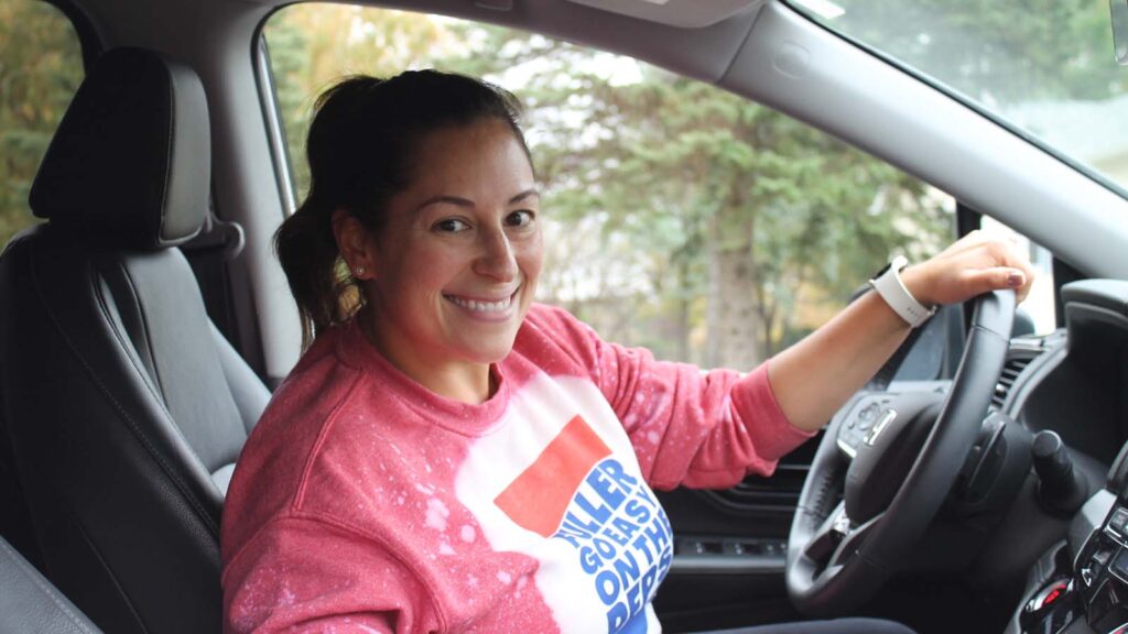 A woman with brown hair smiles with her hand on the steering wheel of her car.