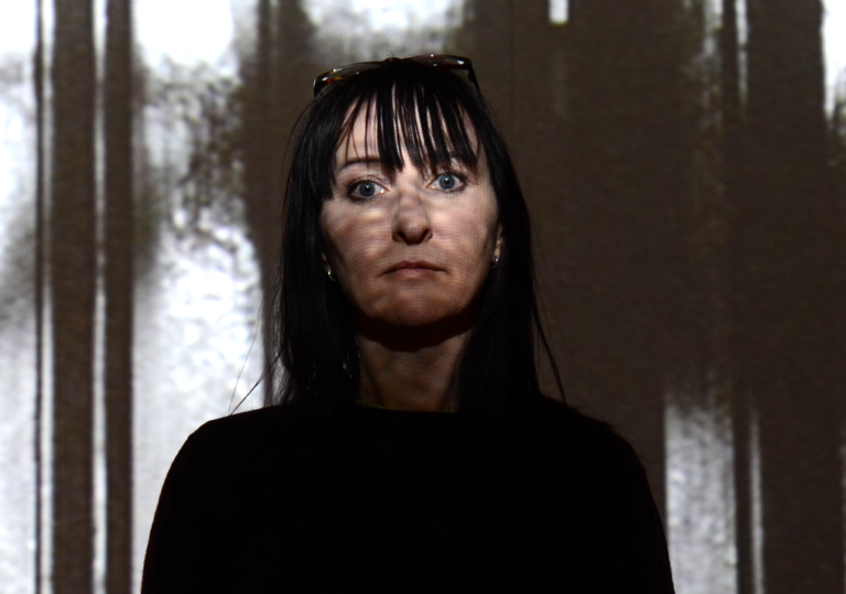 A woman with shadows across her face stands looks into the camera