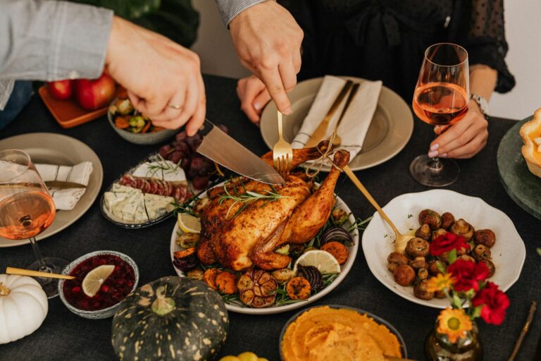 A pair of hands holding a fork and a knife carve a turkey. The turkey is surrounded by other food items like roasted mushrooms, pumpkins and a charcuterie board.