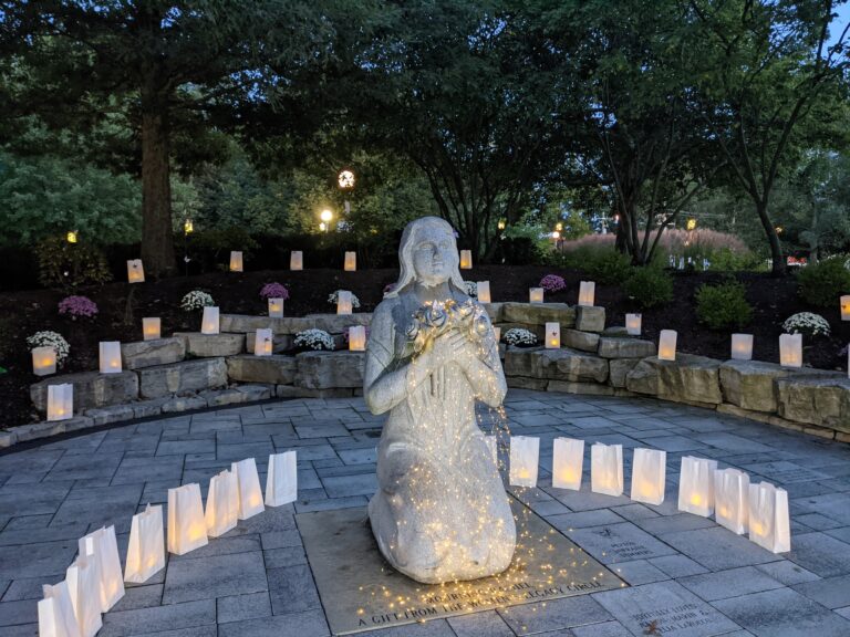 a statue of a kneeling woman, holding flowers in her arms. the statue is covered with lights and surrounded by white paper bags with lights inside.