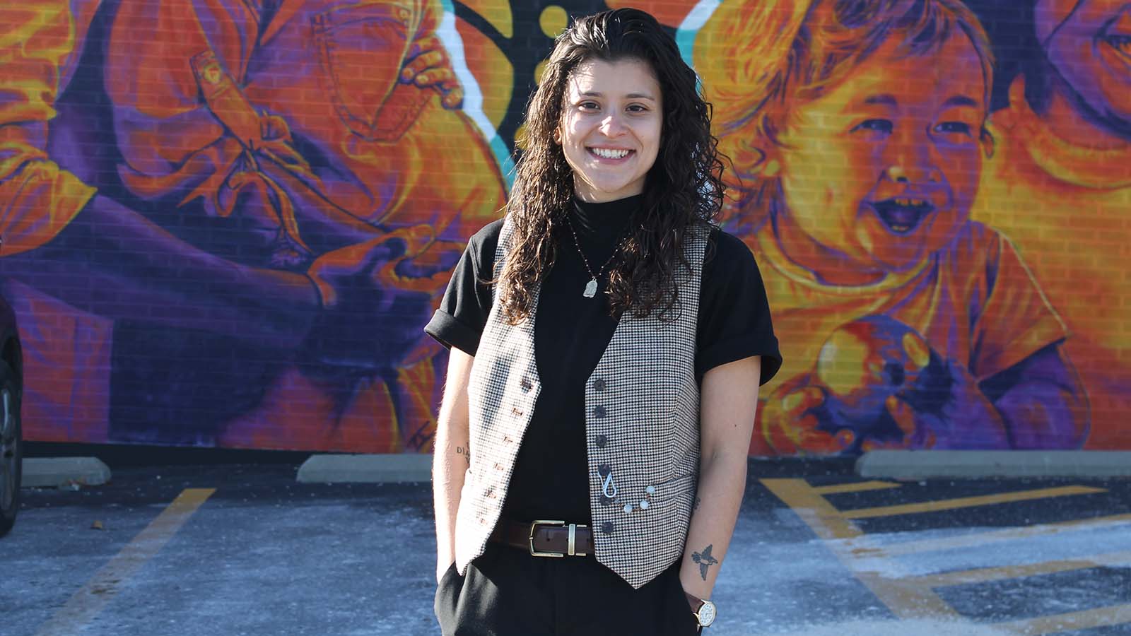 María Jiménez stands with their hands in their pockets in a parking lot with a yellow and purple mural of children and families.