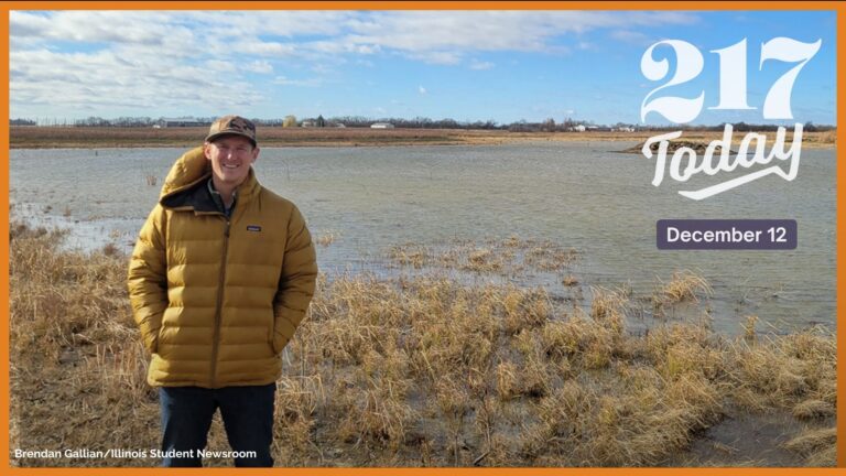 A man in a yellow jacket stands in front of a wetland landscape