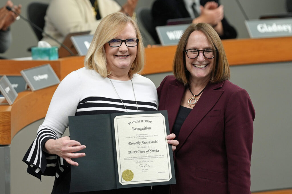 Dorothy David stands in front of members of city council applauding, holding a certificate of recognition.