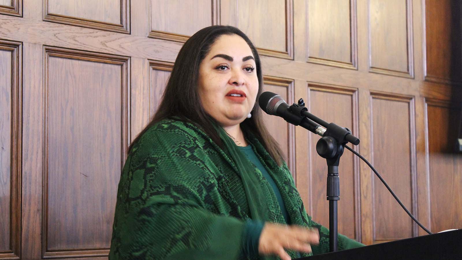 A woman wearing a green jacket and straight brown hair speaks at a podium.