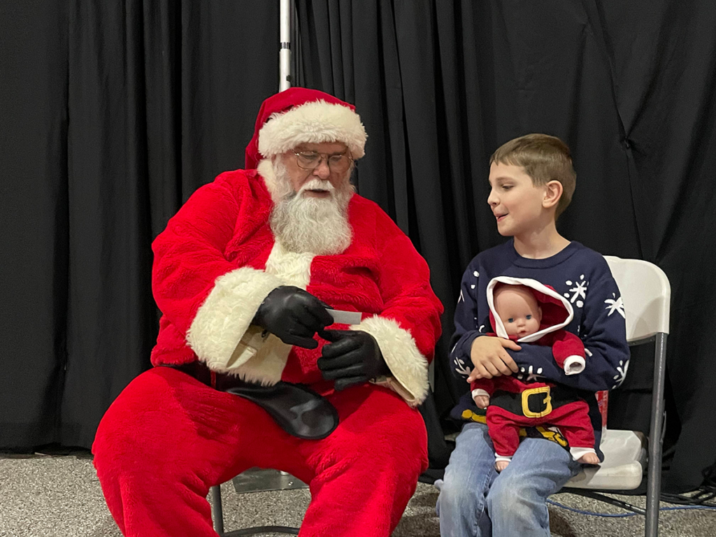 A man dressed as Santa talks to a young boy holding a baby doll.