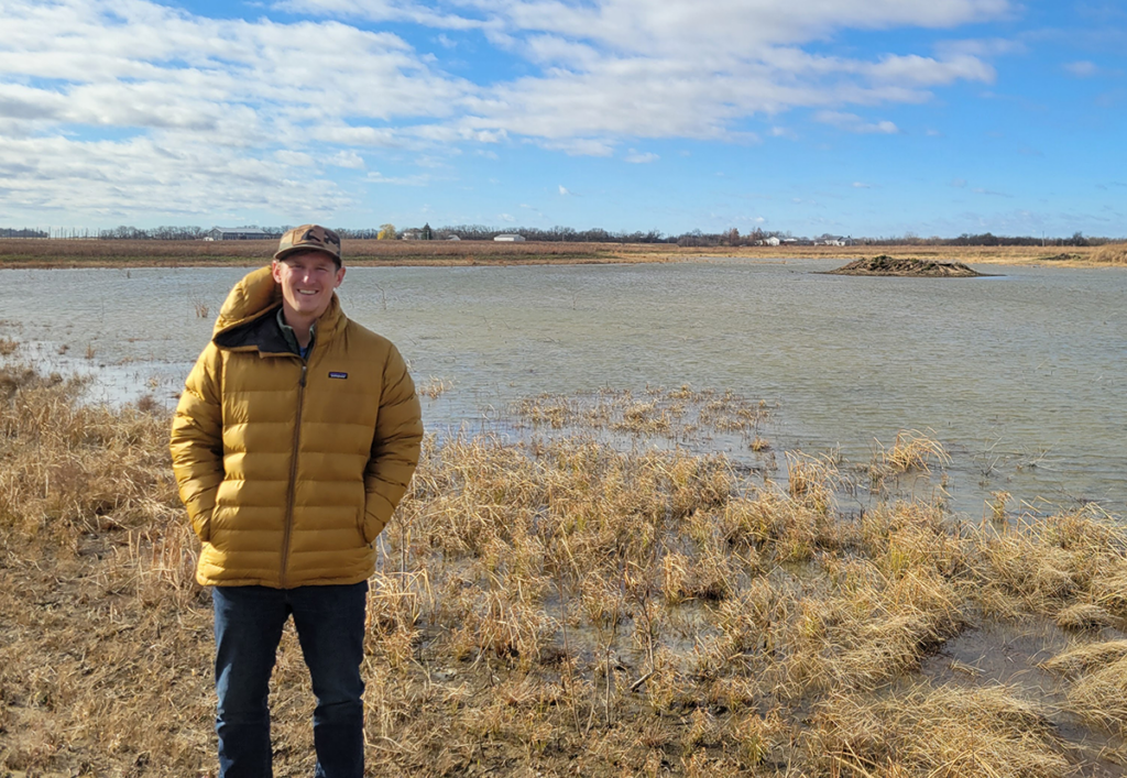 A man in a yellow jacket stands in front of a wetland landscape