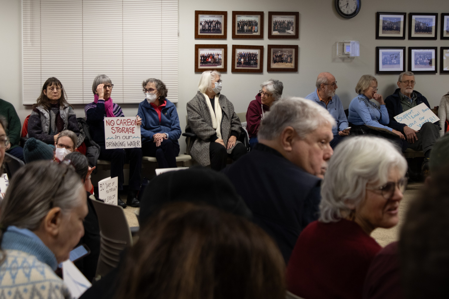 People sit and hold signs that read, "No carbon storage in our drinking water" and "Support local bans. Protect our water."
