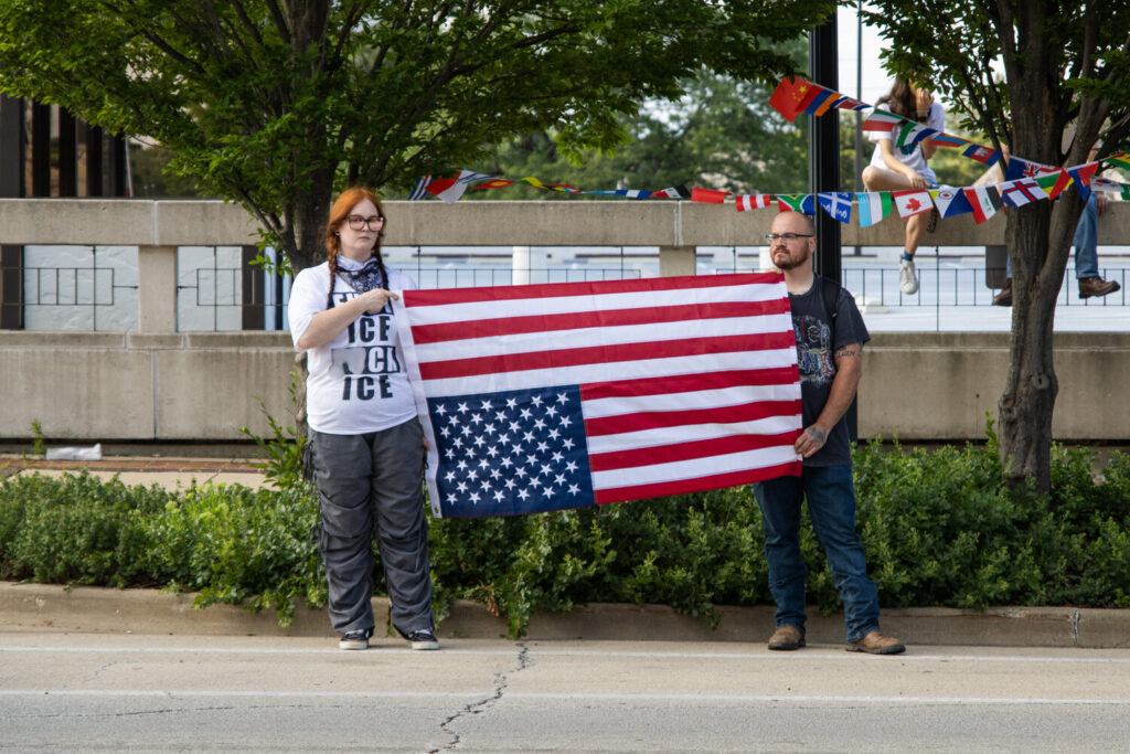 Two people stand across a street, holding up an upside-down American flag between them. Their expressions are neutral.