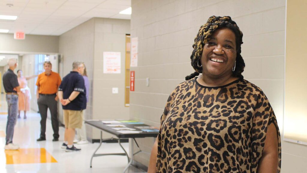 A woman with a leopard print blouse, blonde and brown braids and a big smile stands in the Urbana Sixth Grade Center hallway.