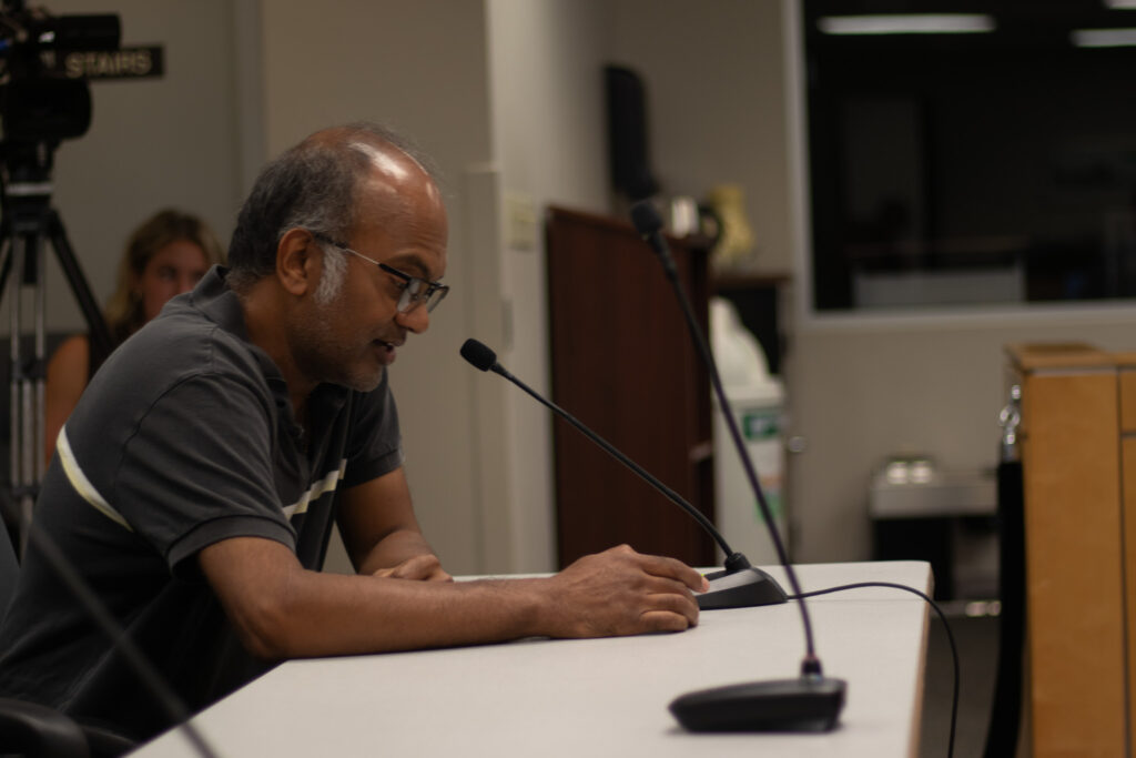A man sits in front of a table and speaks into a microphone.