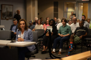 Woman in blue blazer speaking into a microphone in front of a seated crowd.