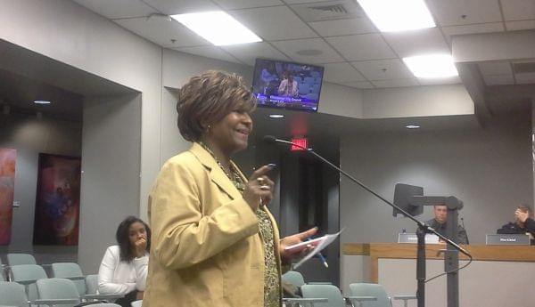 A woman, standing, addresses other at a City Council meeting..
