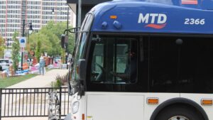 An MTD bus driver stops near the University of Illinois Urbana-Champaign quad in August 2025. The bus takes up most of the frame. A woman in jeans stands in the background further down the road. There are also "Now Leasing" signs and a high-rise apartment in the background.
