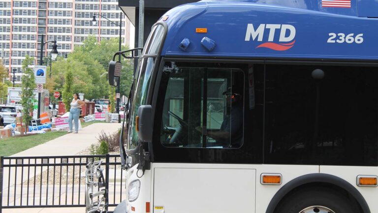 An MTD bus driver stops near the University of Illinois Urbana-Champaign quad in August 2025. The bus takes up most of the frame. A woman in jeans stands in the background further down the road. There are also "Now Leasing" signs and a high-rise apartment in the background.