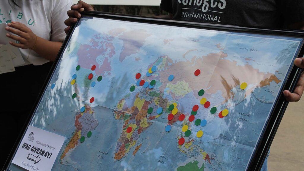 A student holds a world map with colorful dots marking where students come from. Students add dots as they walk by, in mid-August 2025.