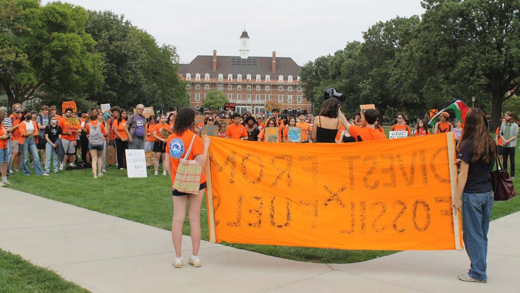 A crowd of students in orange listen to student speakers. In the foreground, two people hold an orange banner. The Illini Union is in the background.