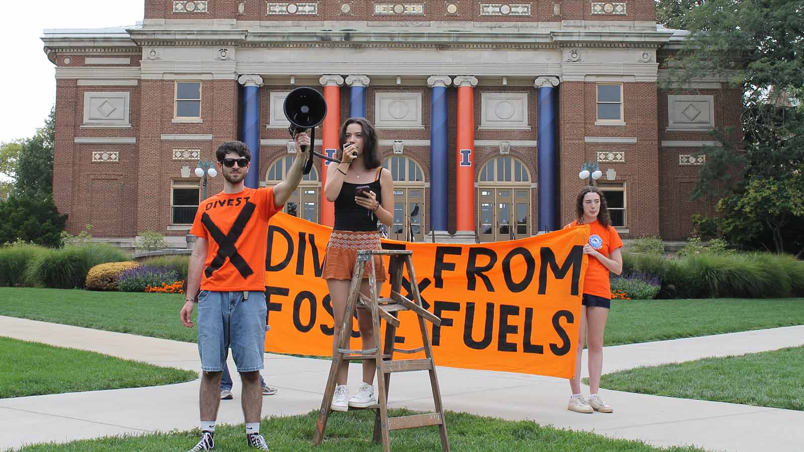 A man wearing an orange shirt with an X on it holds a megaphone for a woman on a ladder on the Quad. Two women hold an orange "Divest from Fossil Fuels" banner behind them. Further in the background are the columns and brick of Follinger Hall.