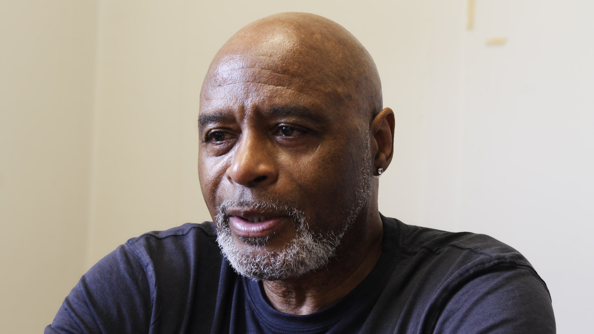 A Black man, who is bald with a short gray beard, stares towards a source of light in his office. His blank, off-white office wall is behind him.