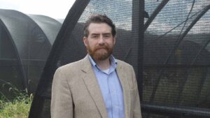 A white man stands in front of a greenhouse-type structure covered in netting.