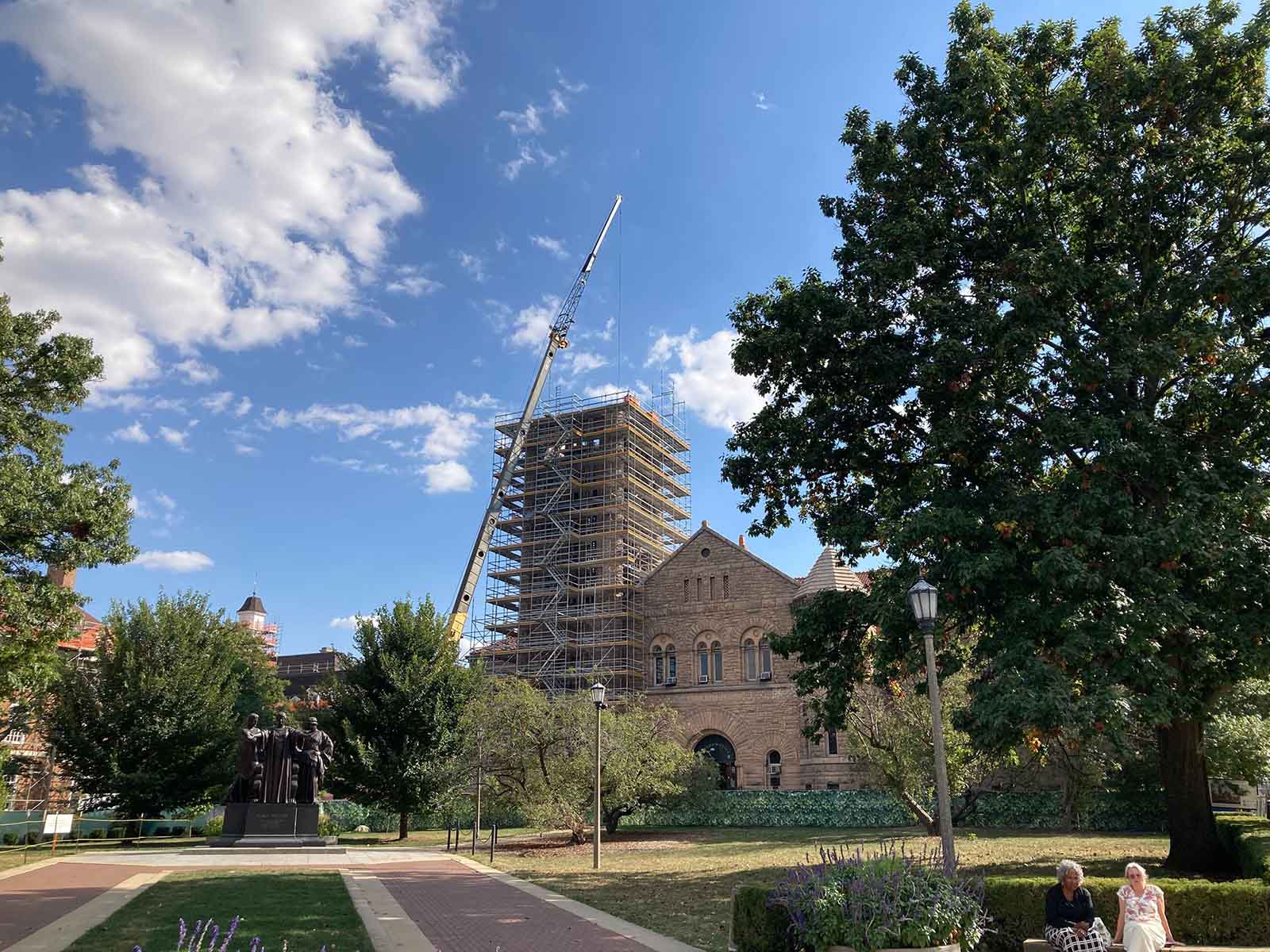 A crane rises above scaffolding on a brick building. The photograph also includes a blue sky, the Alma Mater statue, and two women sitting on a bench.