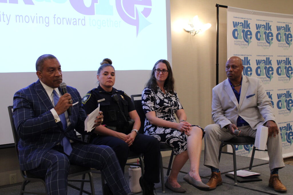 Four people in formal clothing sit in front of a slide show holding microphones.