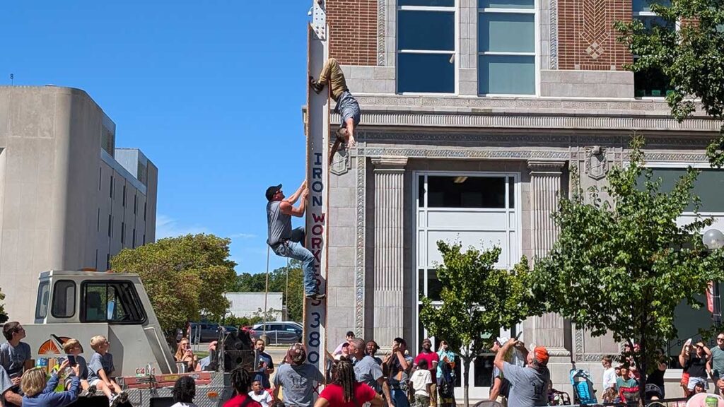 two men climb up a metal beam, one of them hangs upside down hands-free.